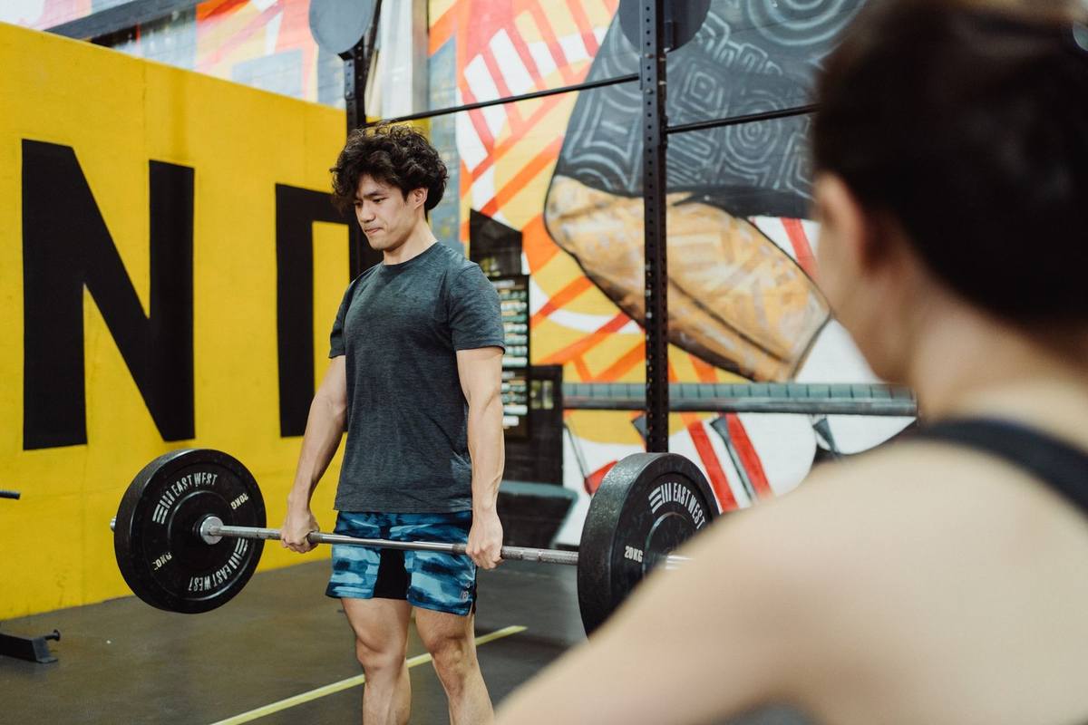 Homme musclé soulevant barrebellée dans salle de musculation avec mur coloré et autre personne en arrière-plan