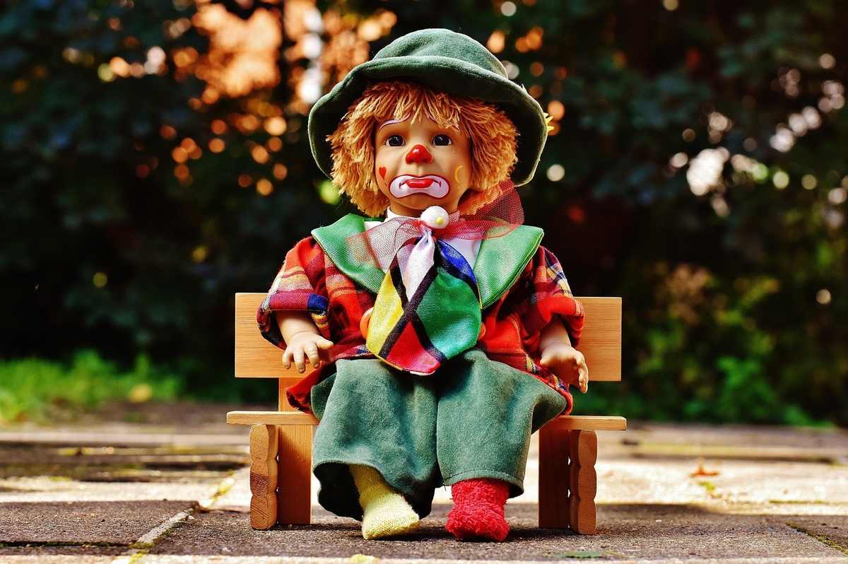 Poupée de clown sur banc miniature, costume coloré, environnement extérieur flou Poupée de clown sur banc miniature, costume coloré, environnement extérieur flou